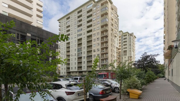 Edificio residencial de varios pisos con balcones, estacionamiento con coches y un sendero bordeado de árboles y arbustos.