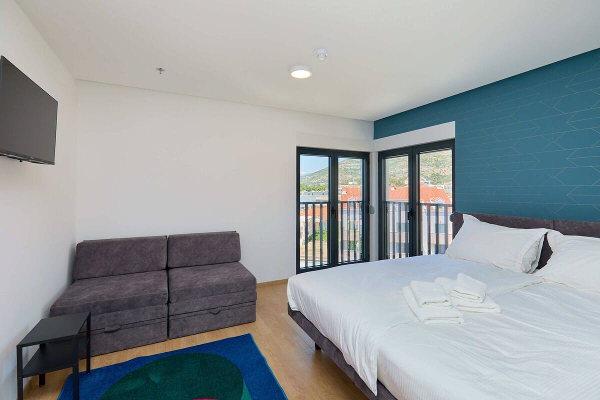 A modern hotel room with a bed, a sofa, a flat-screen TV, and a balcony with a city view.