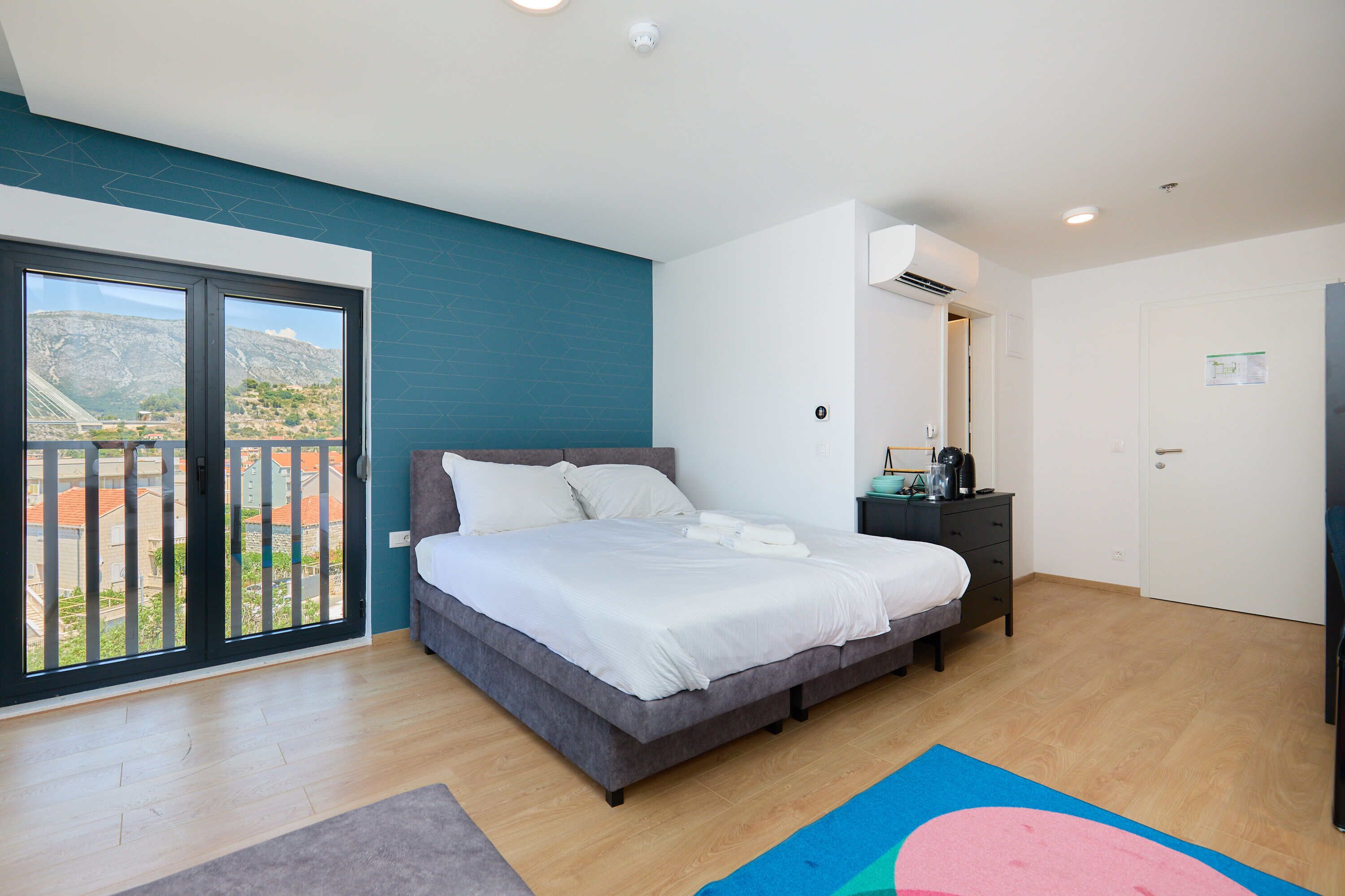 A bedroom with a bed, a nightstand, a dresser, a window with a view, and a door.