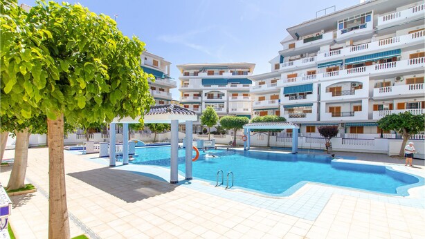 Un área de piscina con una piscina azul cristalina, un deck con baldosas y un edificio de varios pisos con balcones al fondo.