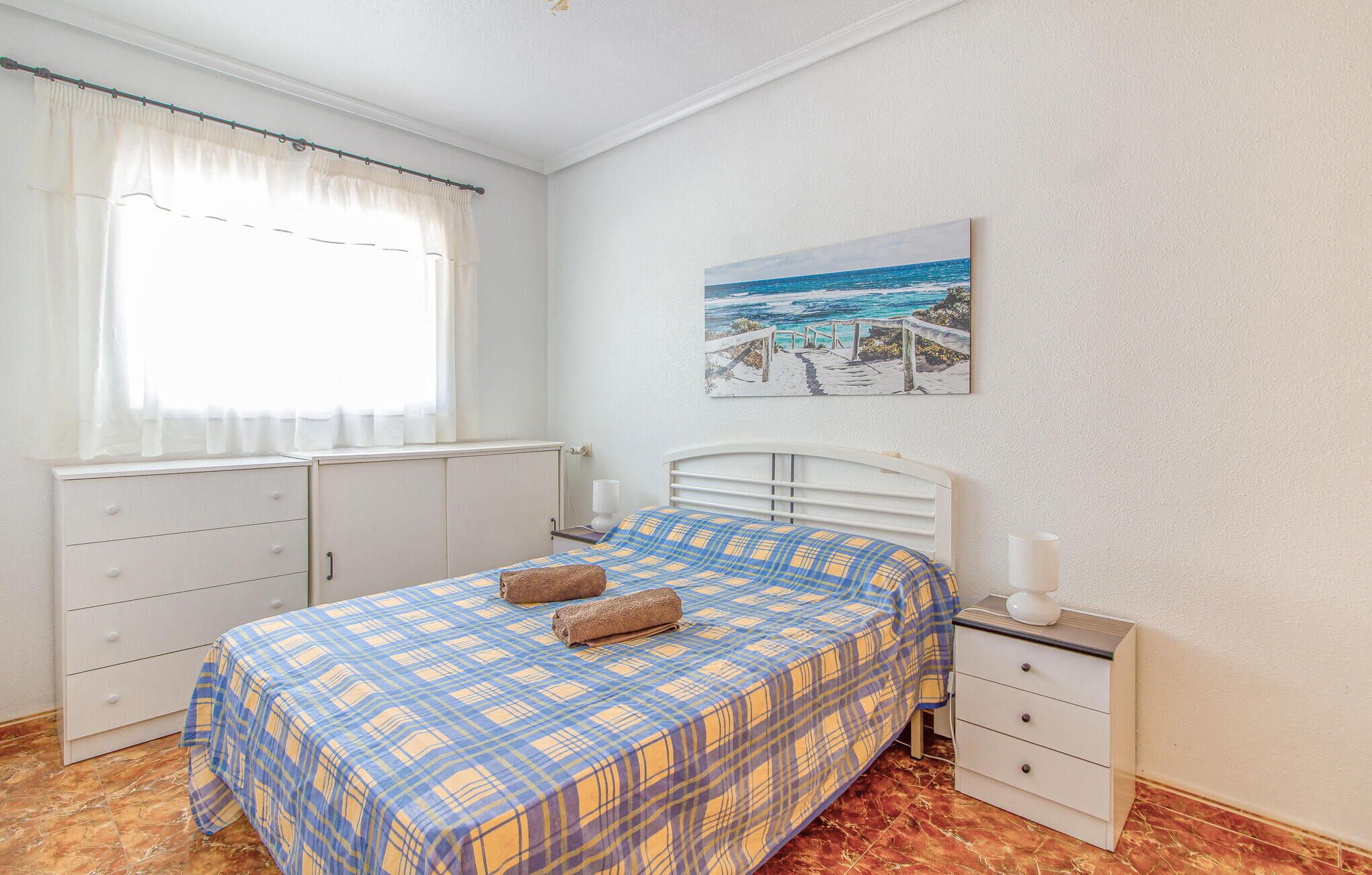 A bedroom with a bed, a nightstand, a window with curtains, a radiator, and a painting of a beach scene.