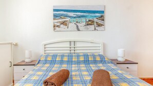 Una cama con colcha a cuadros, dos toallas enrolladas, un cabecero blanco, lámparas de noche y un cuadro de una playa.