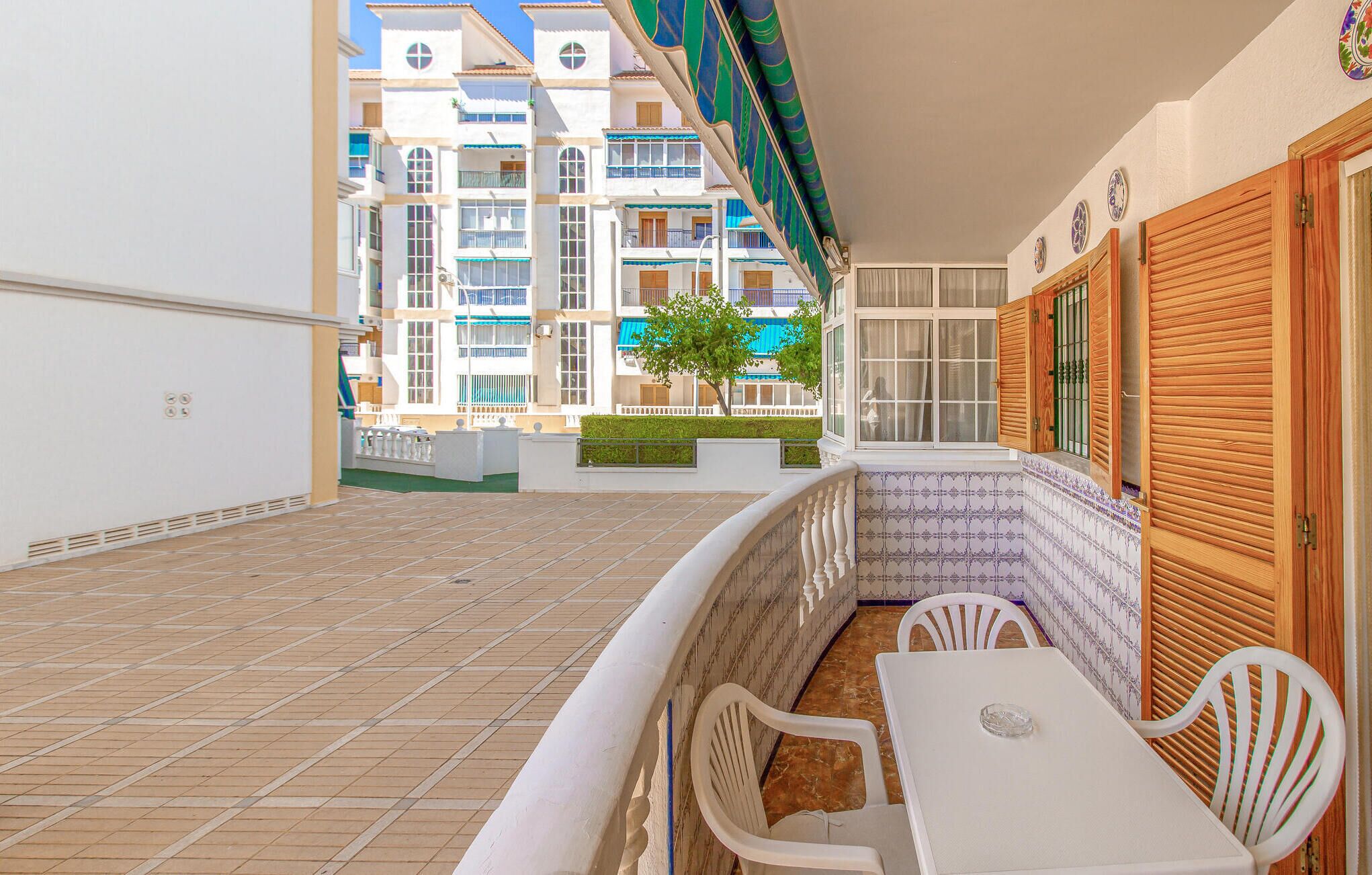 A balcony with a tiled floor, white chairs, a table, and a view of a multi-story building with balconies.