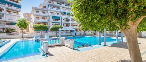 Zona de piscina con suelo de baldosas, un espacio de descanso cubierto y un edificio de varios pisos con balcones al fondo.