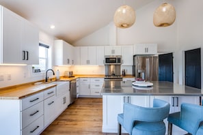 A modern kitchen with white cabinets, stainless steel appliances, and a wooden countertop. There are two hanging light fixtures and a large island with bar stools.