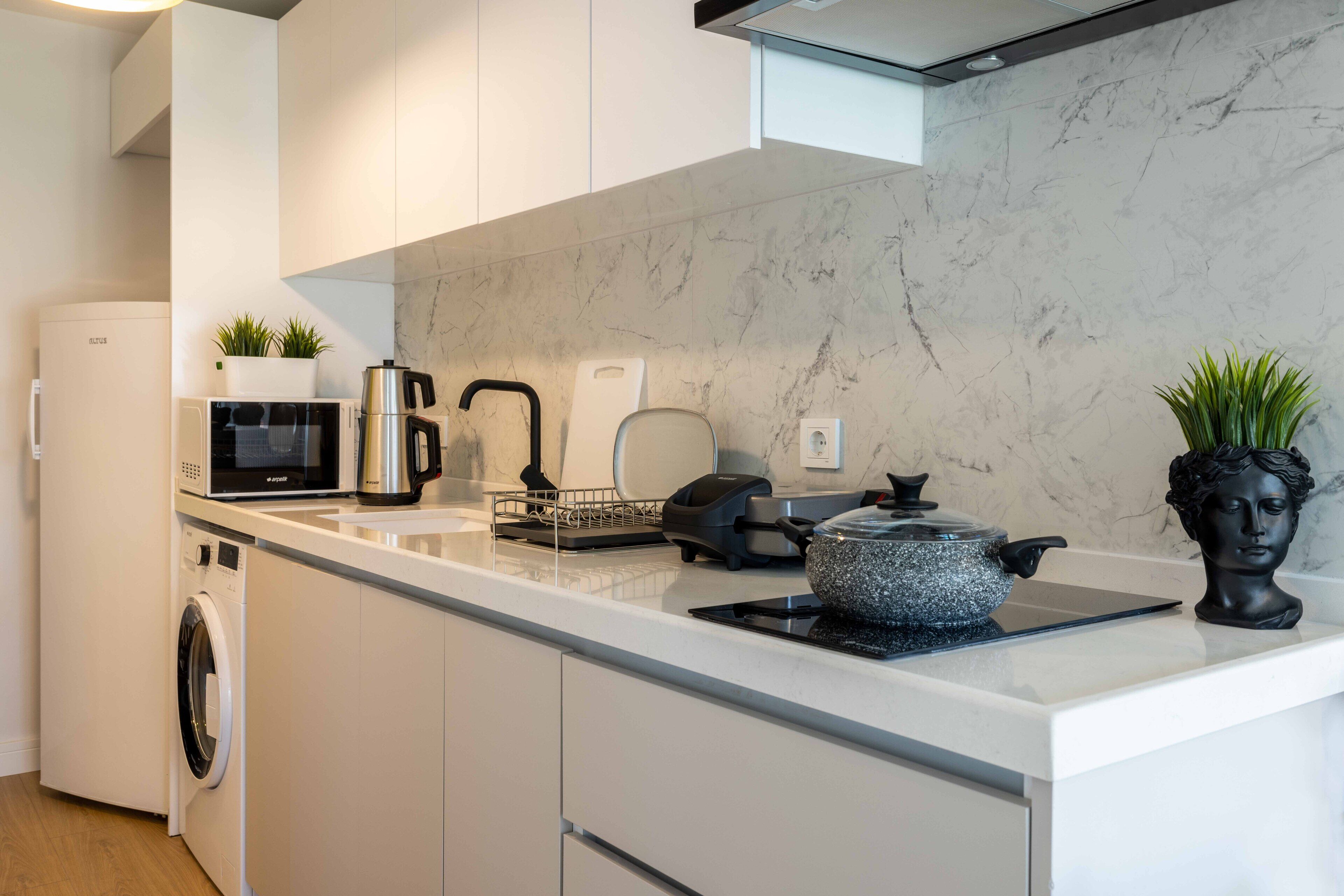 A modern kitchen with a white countertop, a built-in washing machine, a microwave, a kettle, and a decorative bust.