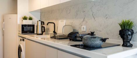 A modern kitchen with a white countertop, a built-in washing machine, a microwave, a kettle, and a decorative bust.