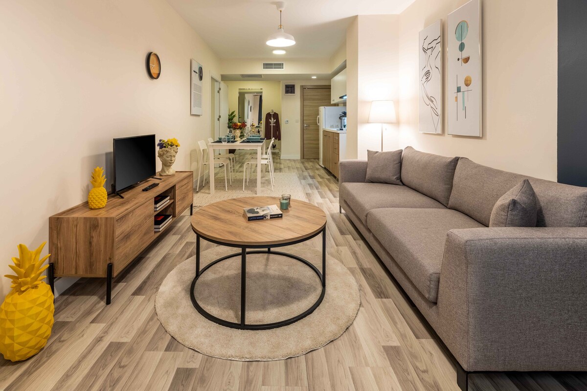 Un salon moderne avec un canapé gris, une table basse en bois et un téléviseur posé sur un meuble en bois. On aperçoit à l’arrière-plan un coin repas avec une table et des chaises.
