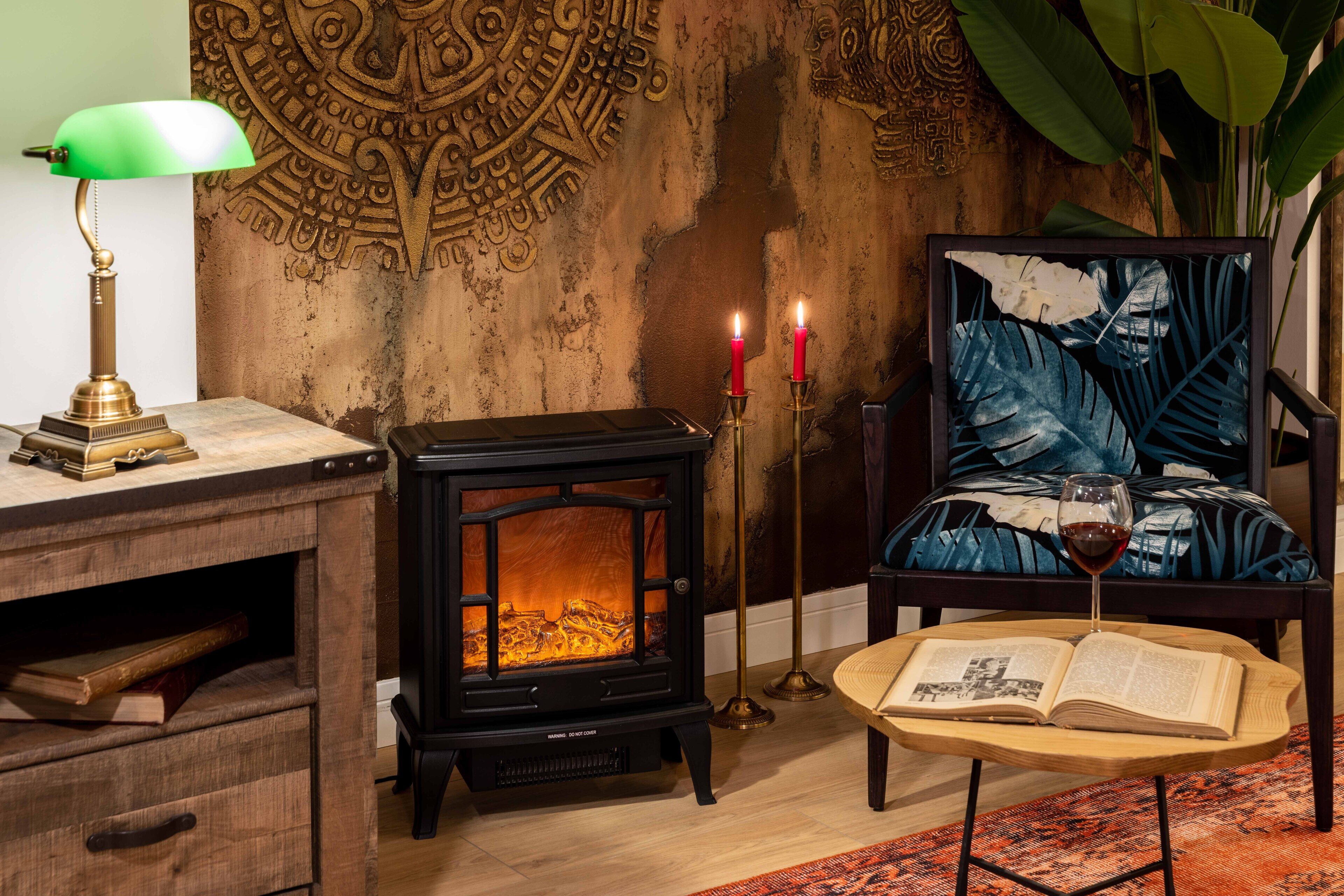 A cozy room with a wood-burning stove, a desk with a lamp, a chair with a book and a glass of wine, and a decorative wall.