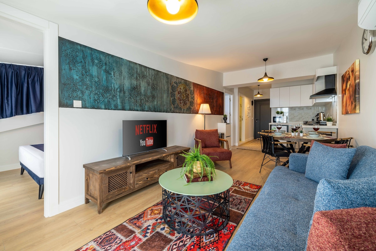 Un salon moderne avec un canapé, une table basse et un téléviseur. On y trouve également un coin repas avec une table et des chaises, et une cuisine à l’arrière-plan.