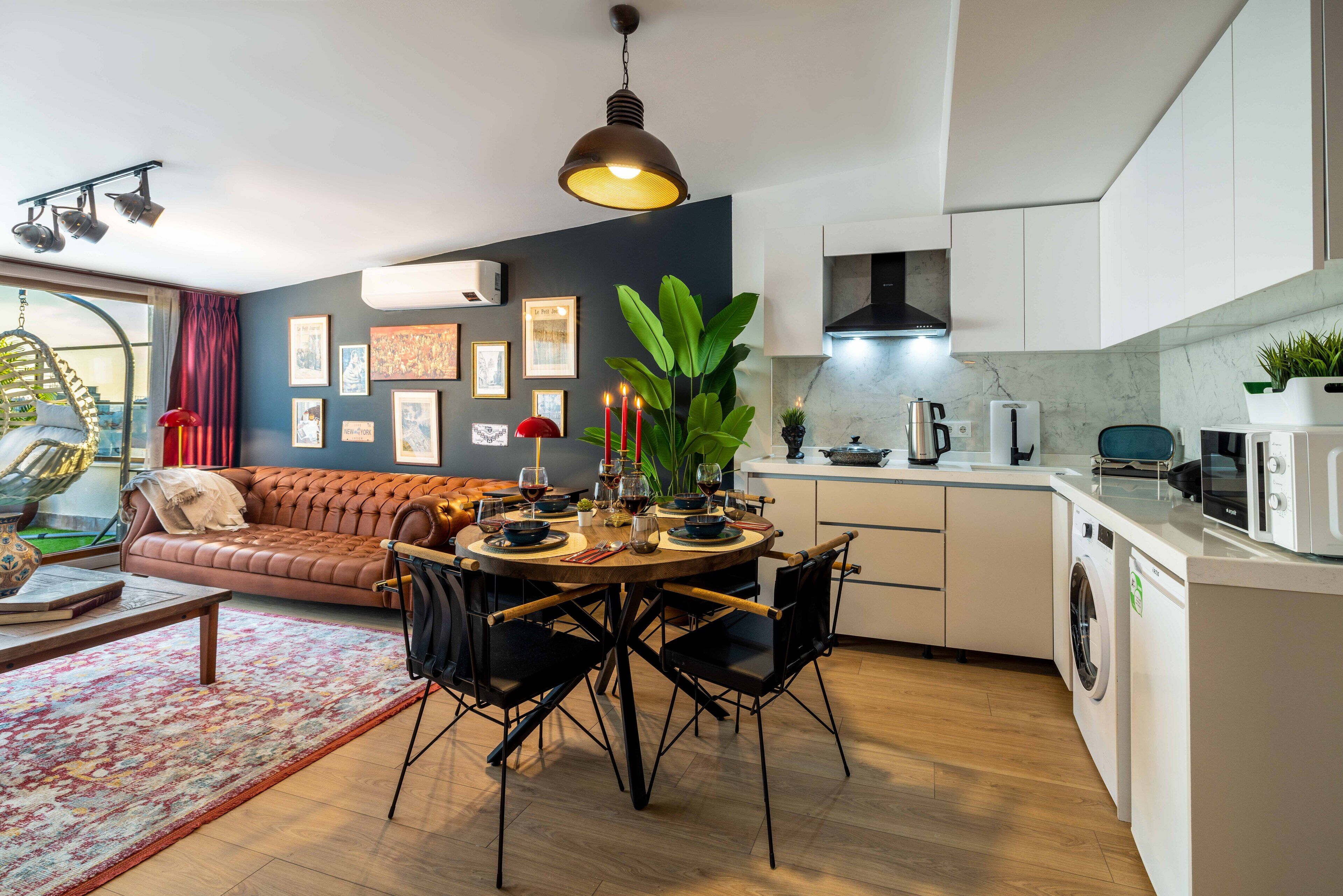 A modern kitchen with a dining area, featuring a round table, chairs, a sofa, and a hanging light fixture.