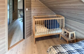 Een houten ledikant met matras en kussens, een klein houten krukje, en een bed met een geruite deken in een kamer met houten wanden en vloeren.