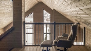Una habitación con techos altos, vigas de madera, un balcón con barandilla y un ventanal con vistas a un paisaje nevado.