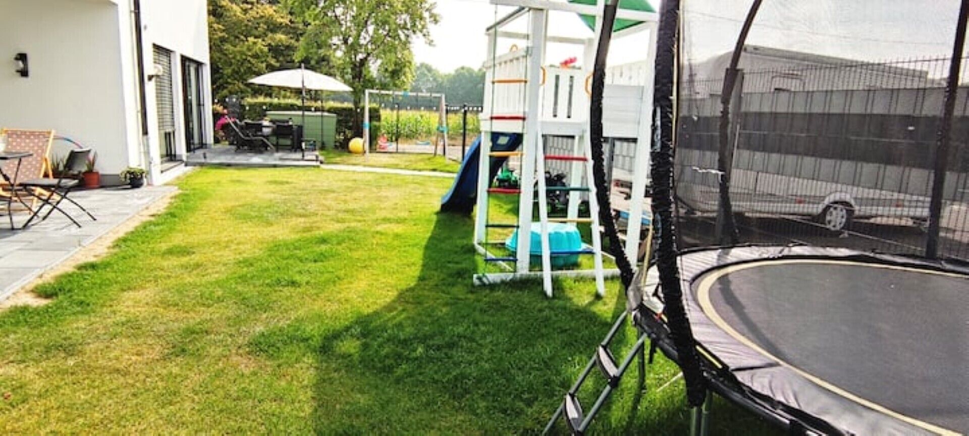 Un jardin avec un trampoline, une aire de jeux et un espace patio aménagé avec du mobilier d’extérieur.