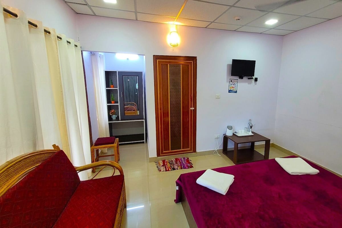 Habitación de hotel con cama, una silla, una mesita, televisor y puerta que da a otra habitación.