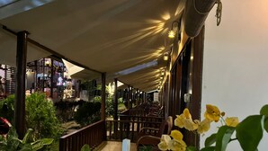 Un área exterior con mesas y sillas, rodeada de plantas en macetas y un paseo techado.