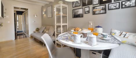 A dining area with a round table, chairs, and breakfast items. There is a sofa and a bookshelf in the background.
