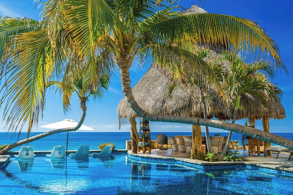 A pool with lounge chairs, palm trees, and a thatched-roof structure.