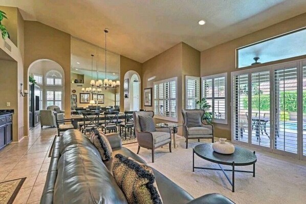 A spacious living area with a leather sofa, armchairs, and a coffee table. There are large windows with blinds, a dining area with a chandelier, and a kitchen visible in the background.