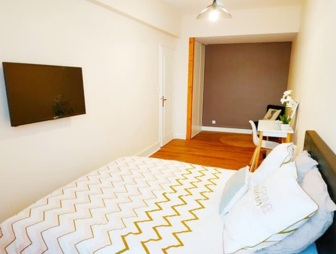 A bedroom with a bed, a white wall, a door, a small table with a plant, and a ceiling light.