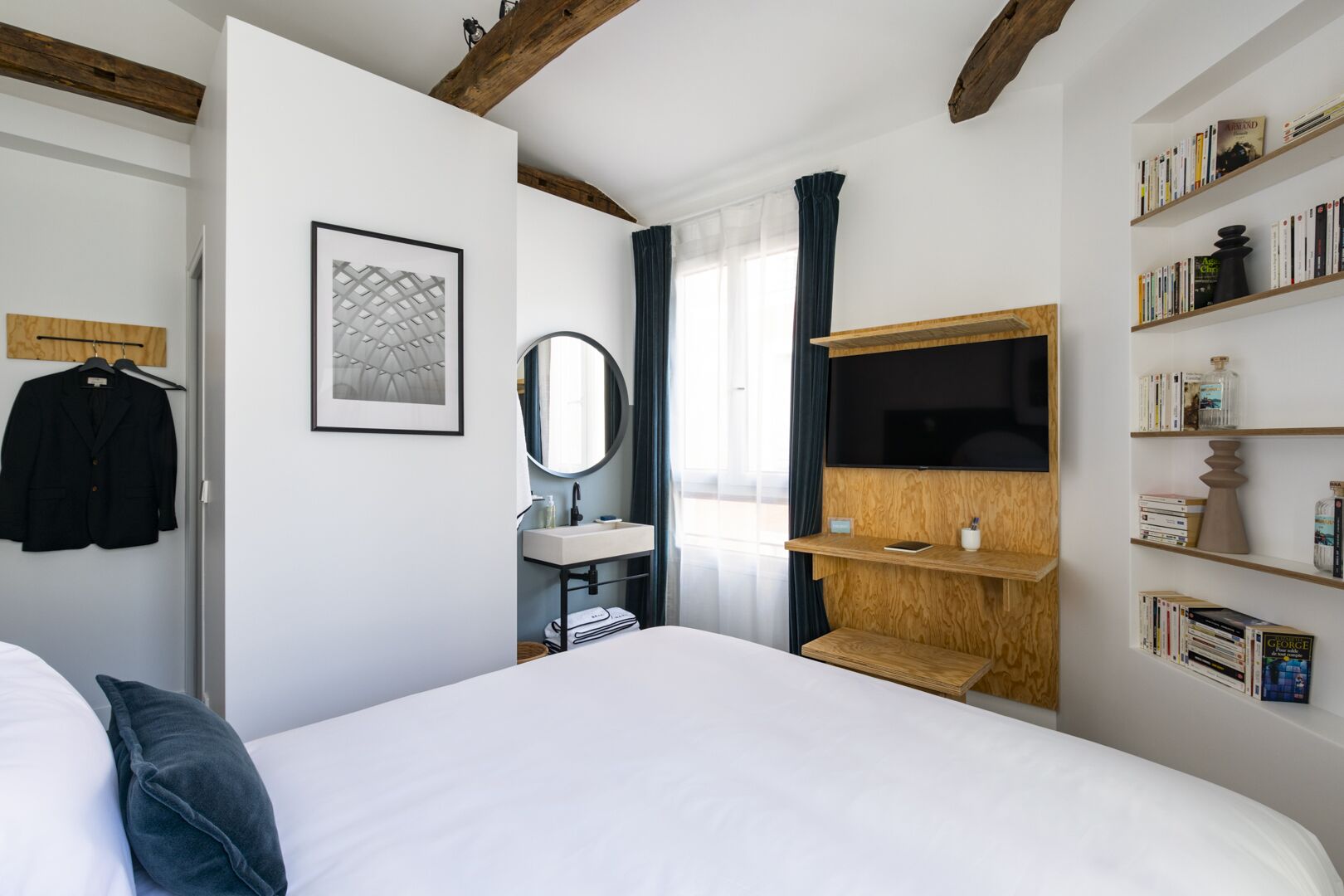 A bedroom with a large bed, a bedside table, a mirror, a TV mounted on a wooden shelf, and a bookshelf filled with books.