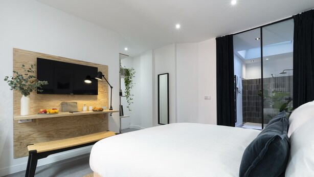Quarto moderno com uma cama grande, televisão montada na parede, mesa de cabeceira com abajur e vista para o banheiro através de uma porta de vidro.
