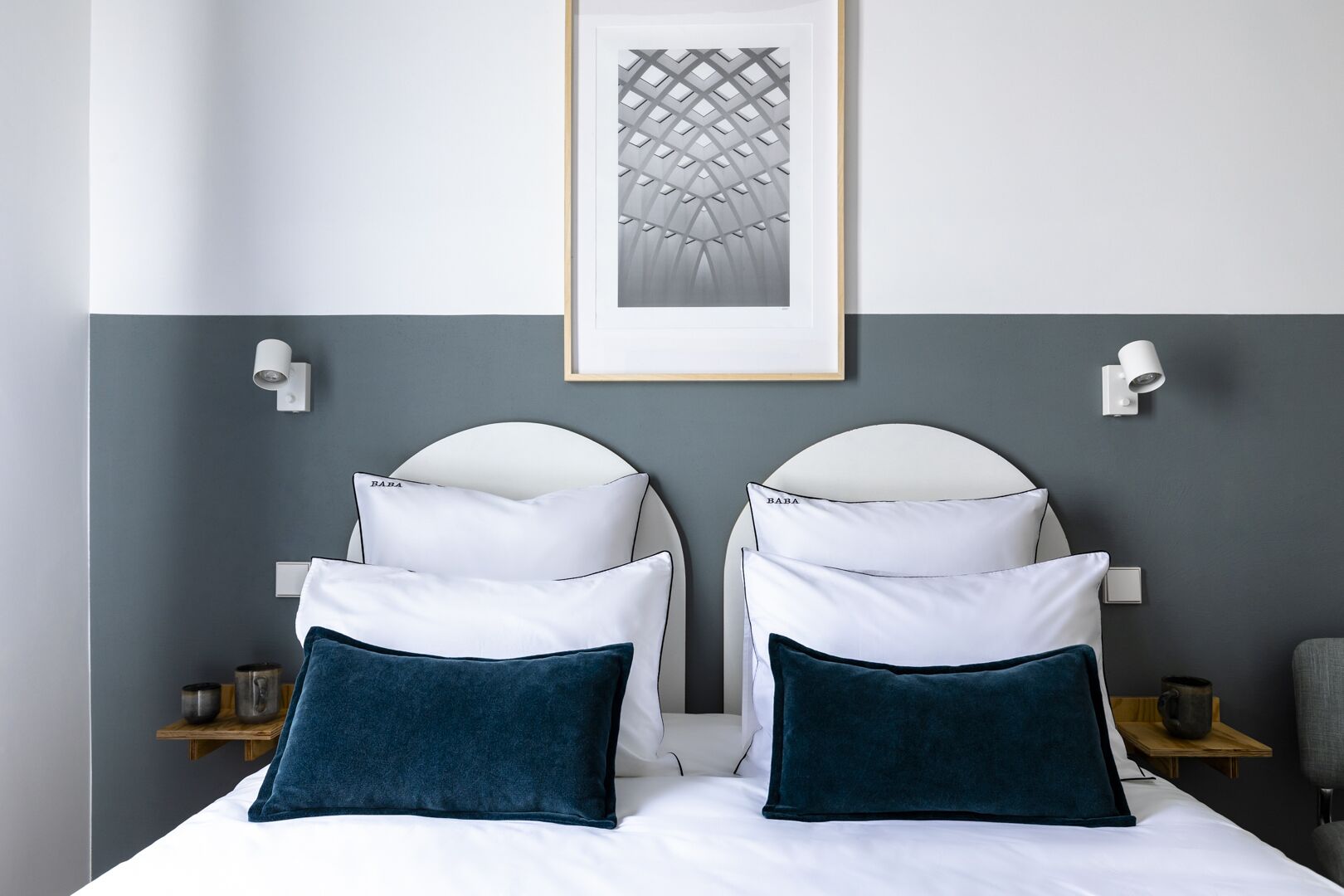 A double bed with white and dark blue pillows, two wall-mounted light fixtures, and a framed abstract artwork above the bed.