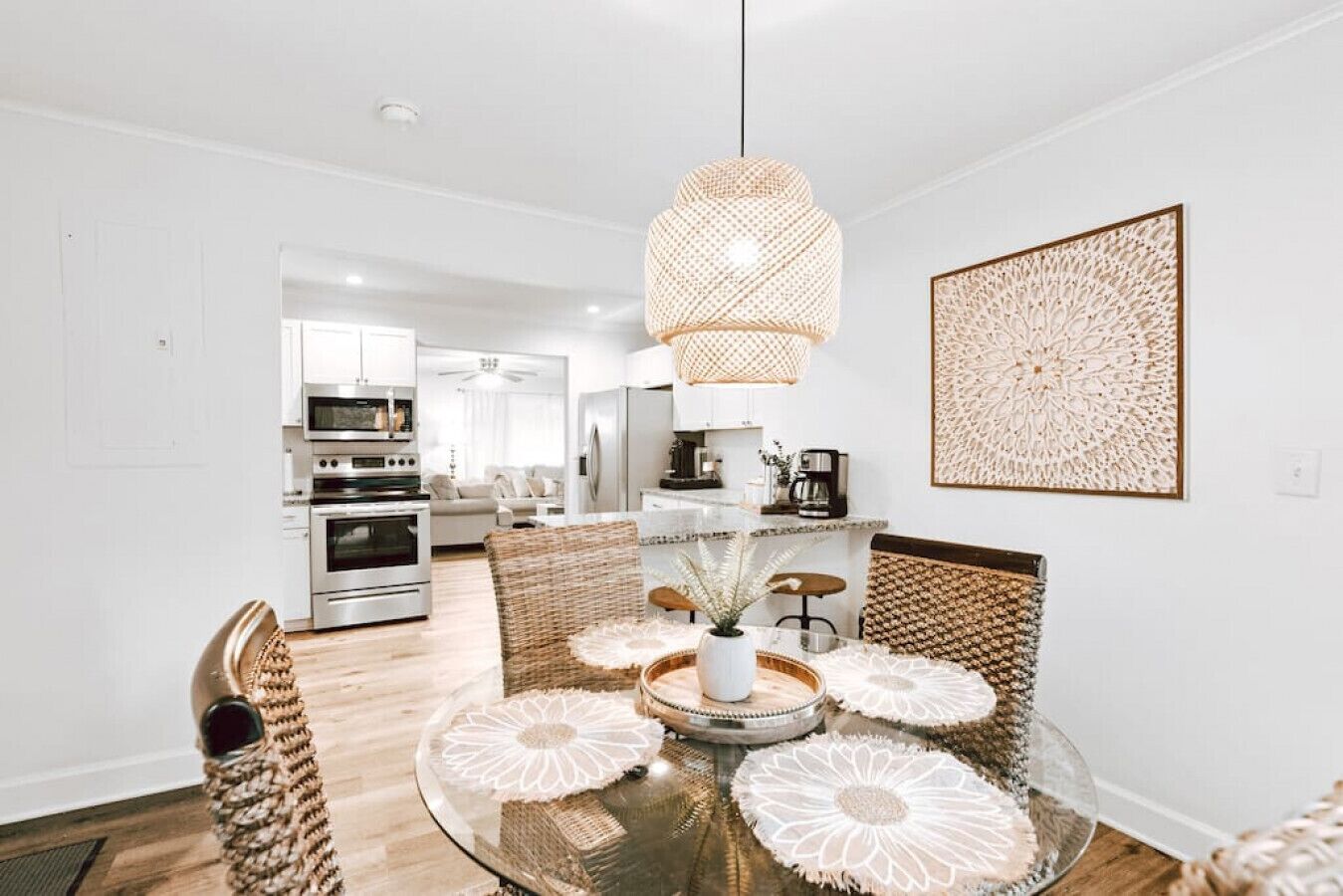 A modern kitchen with stainless steel appliances, a dining area with a glass table and woven chairs, and a decorative wall piece.