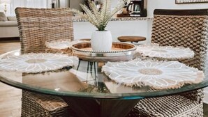 A dining area with a glass table surrounded by wicker chairs, a woven pendant light, and a white vase with a plant.