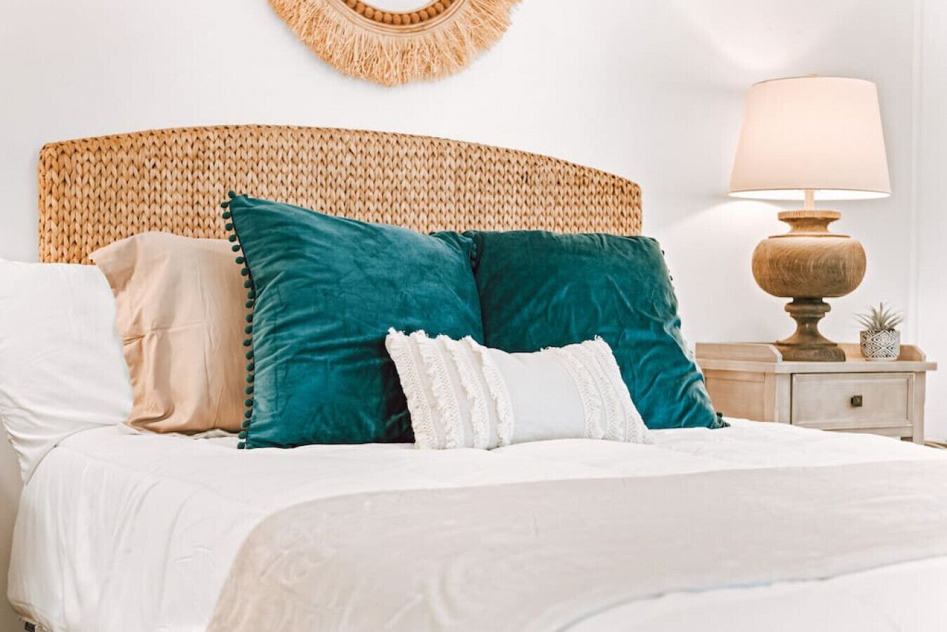 A bed with a woven headboard, multiple pillows, a bedside lamp, and a small potted plant on a nightstand.