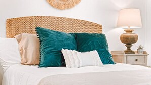 A bed with a woven headboard, multiple pillows, a bedside lamp, and a small potted plant on a nightstand.