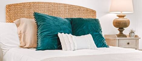 A bed with a woven headboard, multiple pillows, a bedside lamp, and a small potted plant on a nightstand.
