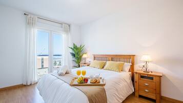 A hotel room with a large bed, a wooden nightstand with a lamp, a tray of breakfast items, and a view of the city through the window.