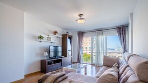 Un soggiorno moderno con televisione a schermo piatto, tavolino e divano. Un balcone offre vista su edifici e alberi.