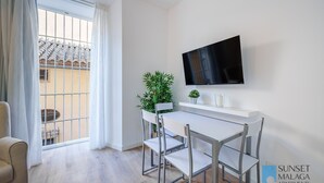En moderne stue med en hvid sofa, et spisebord med stole, et fjernsyn ophængt på væggen og et stort vindue med gardiner.