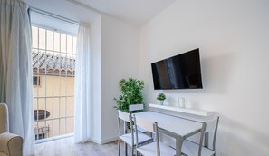 Un saló modern amb un sofà blanc, una taula de dinar amb caires, un televisor penjat a la paret i una finestra gran amb cortines.
