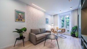 A modern living room with a sofa, a round coffee table, and a dining area with a table and chairs. There are two potted plants, a painting on the wall, and a window with curtains. - Sunset Malaga Apartments - Alameda Apt 1 (Málaga)