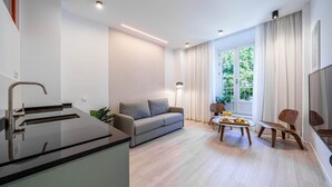 Un salon moderne comprenant un canapé, une table à manger avec un plateau de fruits, un téléviseur et un coin cuisine avec un évier et un robinet.