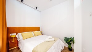 Un lit bien fait, avec une literie blanche et jaune, une table de chevet avec une lampe et une plante en pot dans un coin.