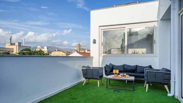 Une terrasse sur le toit avec un mur blanc, un canapé noir, deux fauteuils en osier et une petite table avec des boissons et des en-cas.