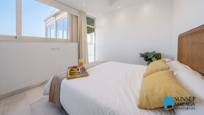 Une chambre avec un grand lit, une table de chevet sur laquelle se trouve une assiette de fruits et un verre de jus d’orange, une plante en pot et une vue sur l’extérieur par une fenêtre.