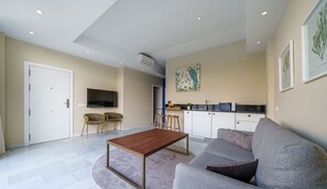Uma sala de estar moderna com um sofá, uma mesa de centro e uma televisão. Há uma área de cozinha com micro-ondas e pia.