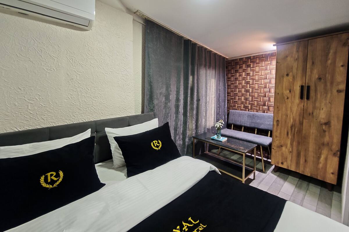 A hotel room with two beds, a wooden wardrobe, a small table, and a grey sofa.