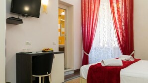 A hotel room with a bed, a desk, a chair, a TV mounted on the wall, and a window with red curtains. - Anthony 47 (Rome)