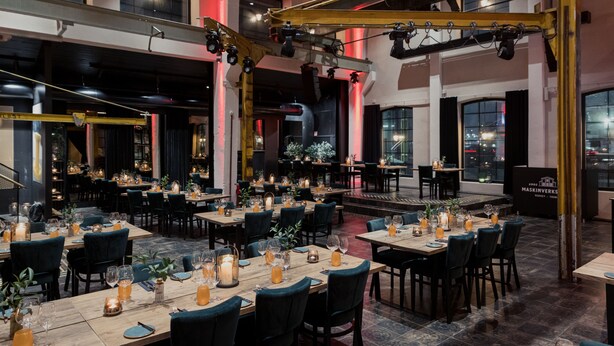 Ein Restaurant mit mehreren gedeckten Tischen, einem Bühnenbereich und großen Fenstern mit Blick auf die Stadt.