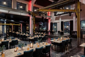 Un restaurante con varias mesas dispuestas para comer, que cuenta con un escenario y amplios ventanales con vistas a la ciudad.