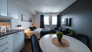 Ein modernes, kompaktes Hotelzimmer mit einer Küchenzeile, einem Bett mit dunkler Bettwäsche, einem an der Wand befestigten Flachbildfernseher und einem Essbereich mit Tisch und Stühlen.