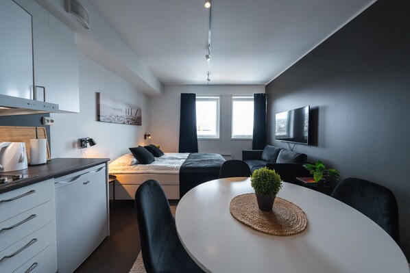 A modern, compact hotel room with a kitchenette, a bed with dark bedding, a flat-screen TV mounted on the wall, and a dining area with a table and chairs.