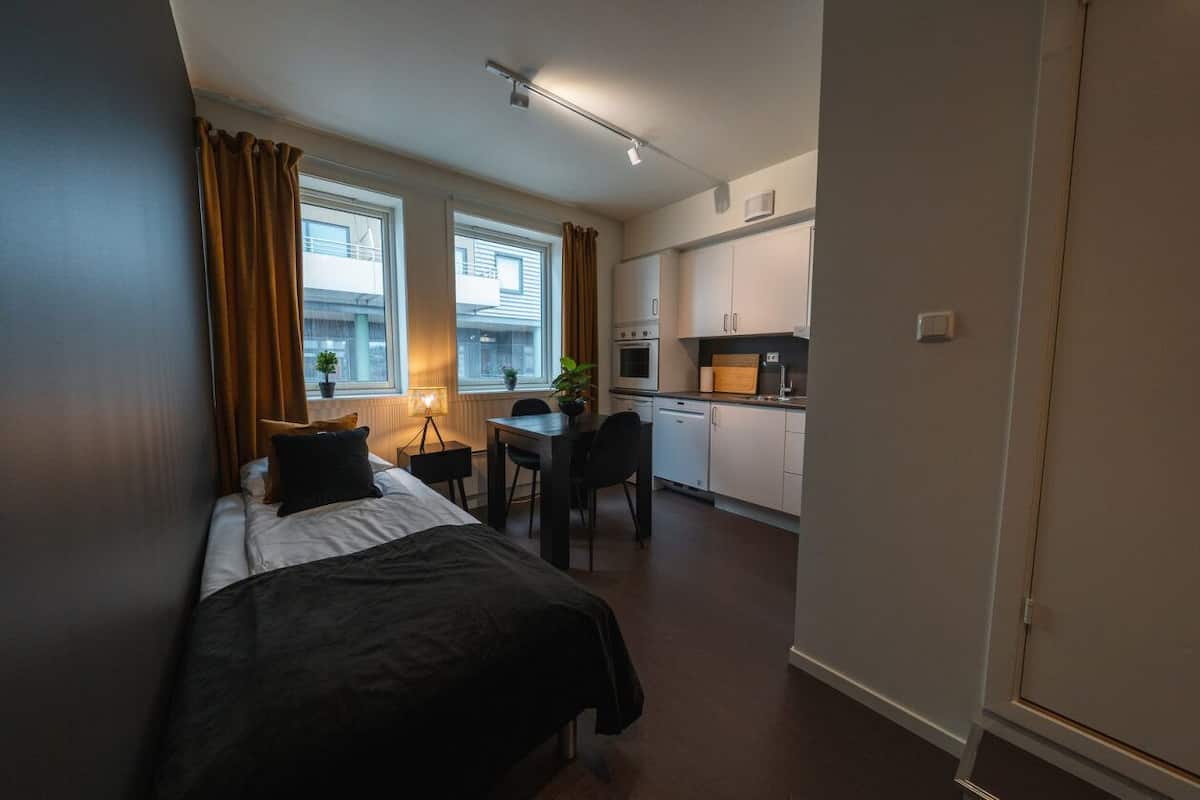 A compact bedroom with a bed, bedside table, and a small desk area with a chair. A window with curtains and a plant is visible.