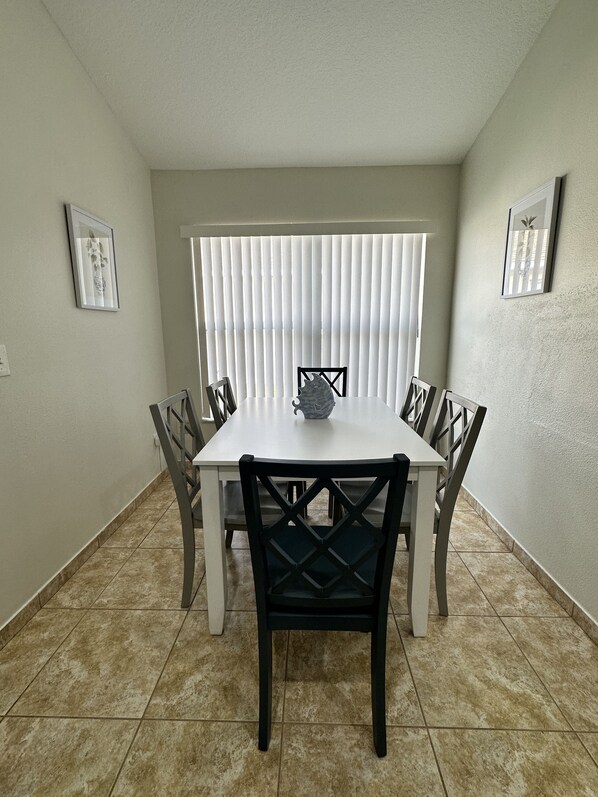 A dining area with a white table and black chairs, a window with blinds, and a tiled floor.